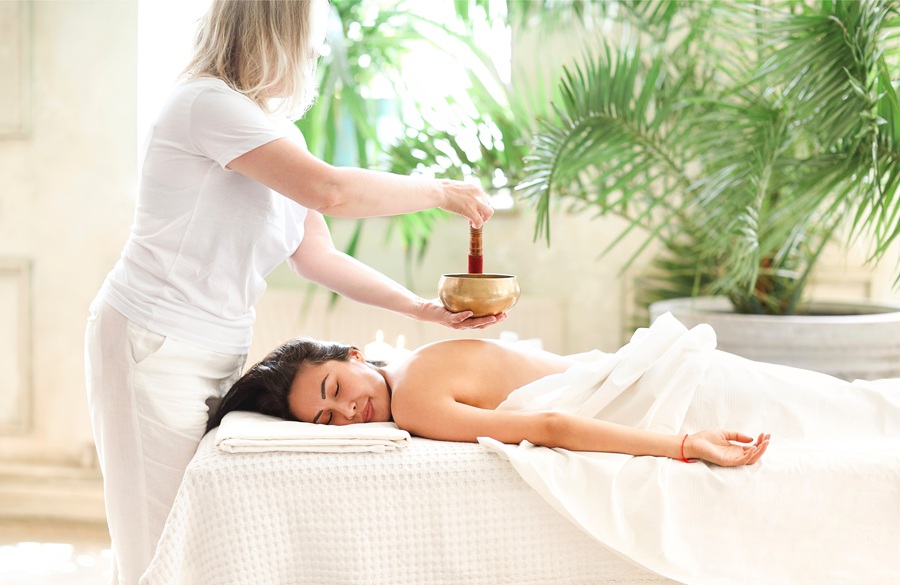 Side view of female therapist using singing bowl over back of relaxed Asian woman in spa salon